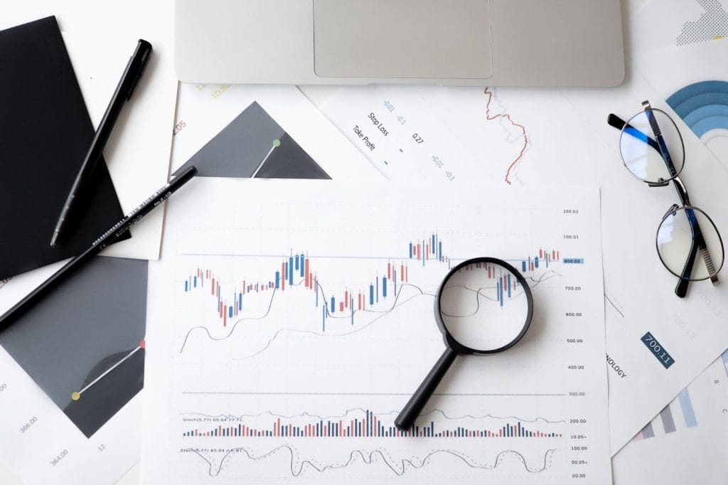 Magnifying glass on financial candlestick charts with eyeglasses, pens, and laptop on a white desk.