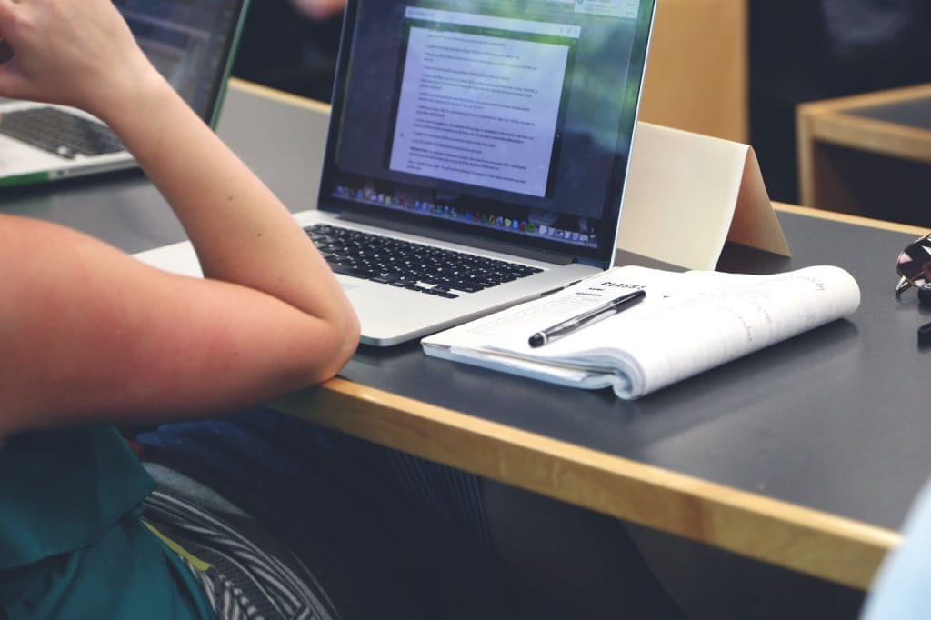 Person typing on laptop next to an open notebook and a pen in a classroom or office setting.