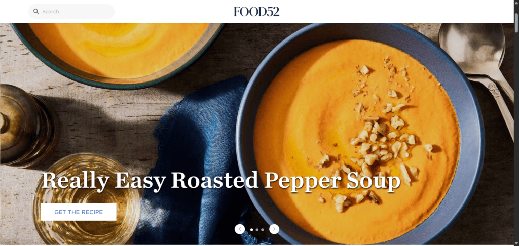 Bowl of roasted pepper soup topped with walnuts next to a wine glass and napkin on a wooden surface posted on the Food52 website