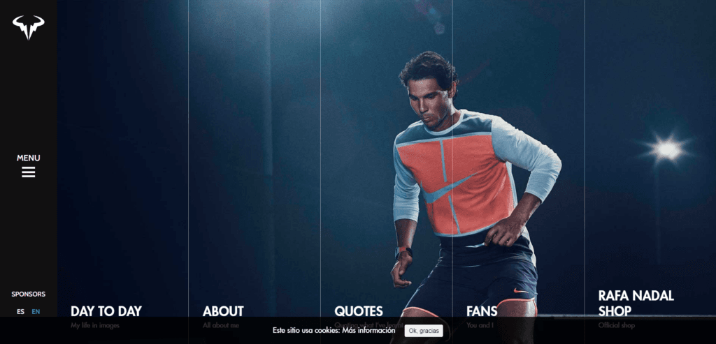 A screenshot of Rafael Nadal's official website featuring a dark, high-contrast design. The background shows a professional photograph of Nadal in his tennis gear under a spotlight. A vertical black sidebar on the left contains the main menu and language toggles for Spanish and English. The bottom of the screen displays large, white navigation links for "DAY TO DAY," "ABOUT," "QUOTES," "FANS," and "RAFA NADAL SHOP," each with a brief subtitle in grey text. A small cookie consent banner in Spanish is visible at the very bottom center.